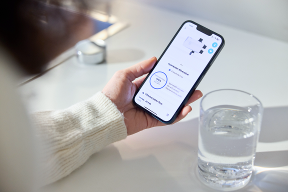 Eine Person hält ein Smartphone mit einer App, daneben ein Glas Wasser auf einem Tisch.